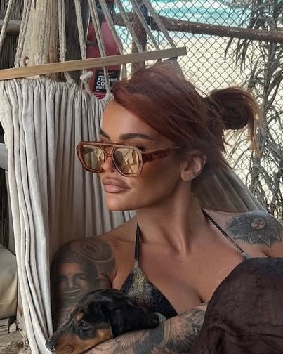 Woman with tattoos wearing sunglasses and a black top, sitting on a hammock with a dog.